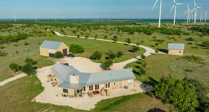 Ranch home in foreground with wind turbines and shrubs in background on green field