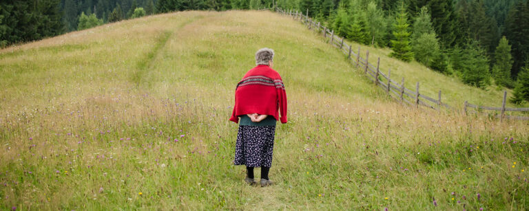 the back of an elderly woman with red coat draped over her shoulders looking over a grass covered hillside with fence and trees contemplating