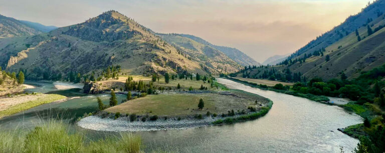 wide river meanders around the tip of a wilderness area with mountains, tree and shrubs scattered throughout