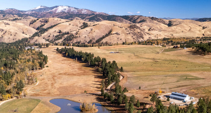 high overview of ranch with barn and pond in foreground and tree-lined creek snaking through pasture to distant hills and then mountains