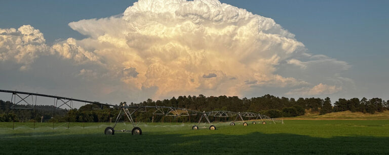 high piled dense clouds rise above a tree covered hill with pivot on green field in foreground