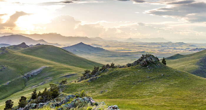 drifting clouds cover distant sloped mountains with green covered slopes in the foreground, piles of rock and pockets of junipers