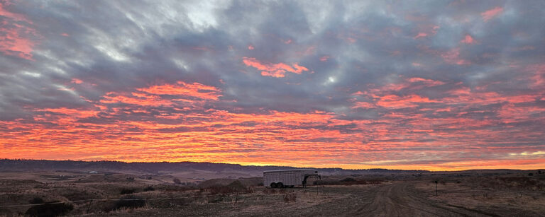 gooseneck horse trailer sits in mountain pasture with setting sun lighting up the clouds in pink and yellow