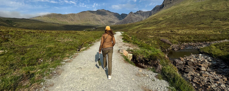 back of woman hiking up well-traveled gravel trail holding small pack walking towards distant mountains