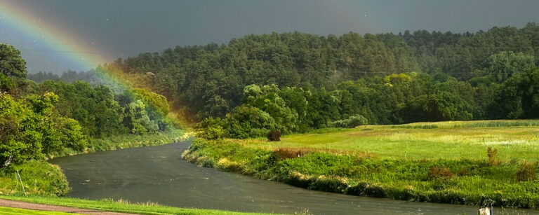 rainbow over a bend in the river wrapping around a green pasture with hills in background