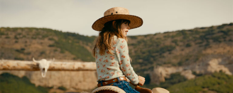 young girl in wide-brimmed straw hat horseback with treed hills in the background