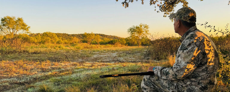 man in camouflage sitting on stump with rifle across his knees watching an open field
