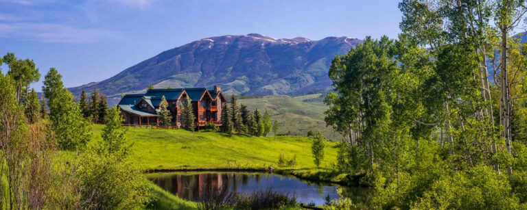 large log home sits on hill above pond with rising mountains as a backdrop