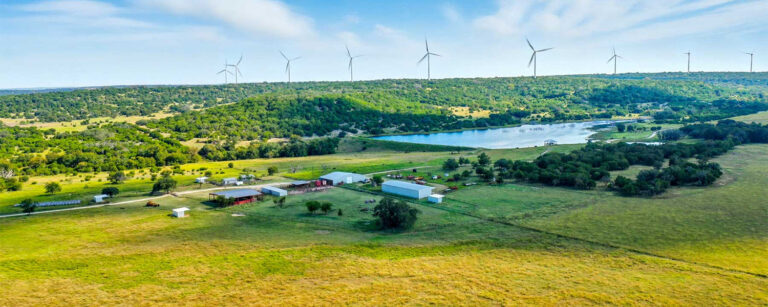 green pasture with buildings, a lake, and wind turbines on a hill.