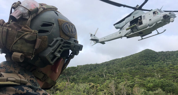Man in Marine fatigues with helmet watches a helicopter landing in field