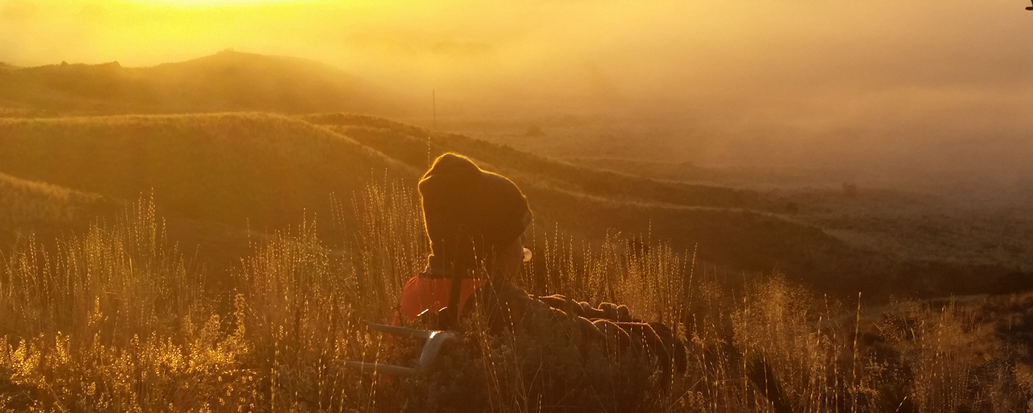 person sitting on hillside in camo, blowing bubble gum and looking out over orange sunlit clouds and mountain valley