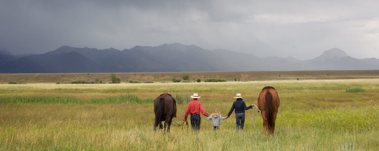 Two adults walking and leading horses with child in between them on grass meadow with cloudy sky and fog covering distant mountains