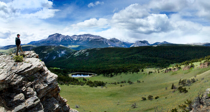 man perched on rocky outcropping overlooking green valley with pond and bordered behind by dense evergreen forest with dark blue mountains and blue sky with cumulus clouds peppered on it